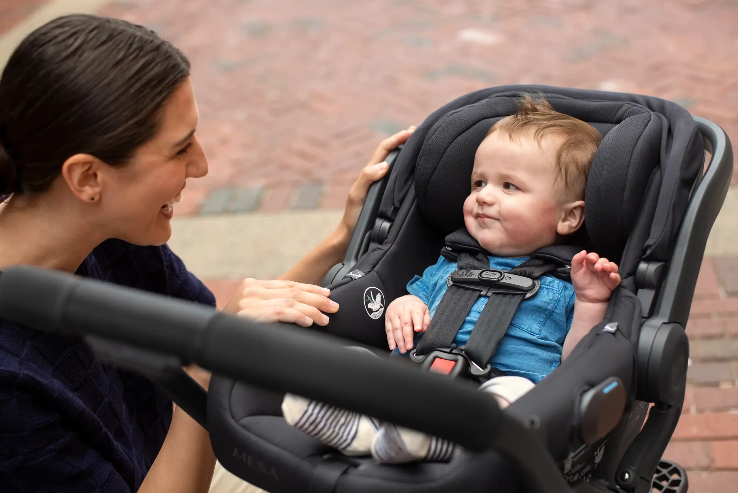 A smiling woman looks at her child securely buckled in the Mesa V2 with its one crotch buckle position and no-rethread, adjustable harness