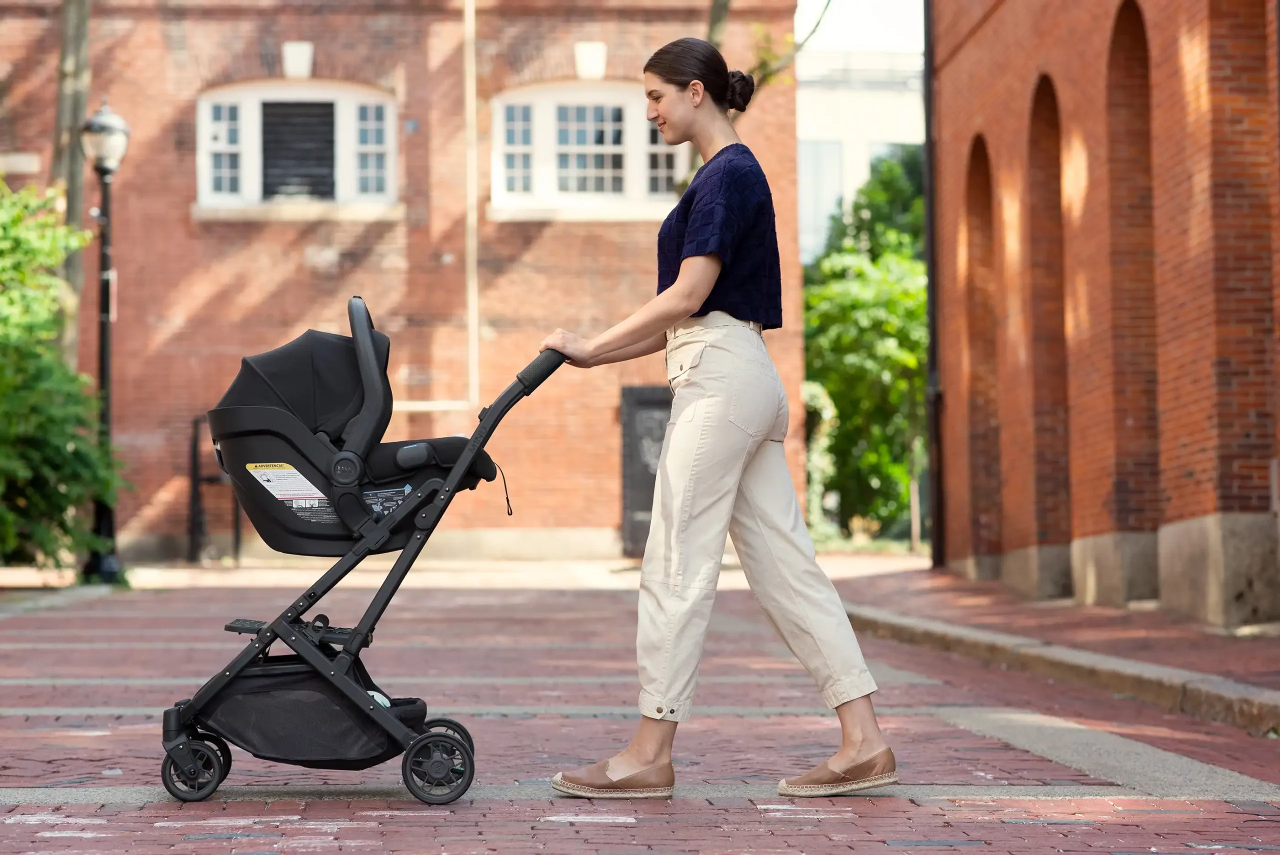 A smiling woman strolls with her child in the Mesa V2 infant carrier attached to the Minu V2 travel stroller with the use of convenient adapters that allow her to go from drive to stroll with ease