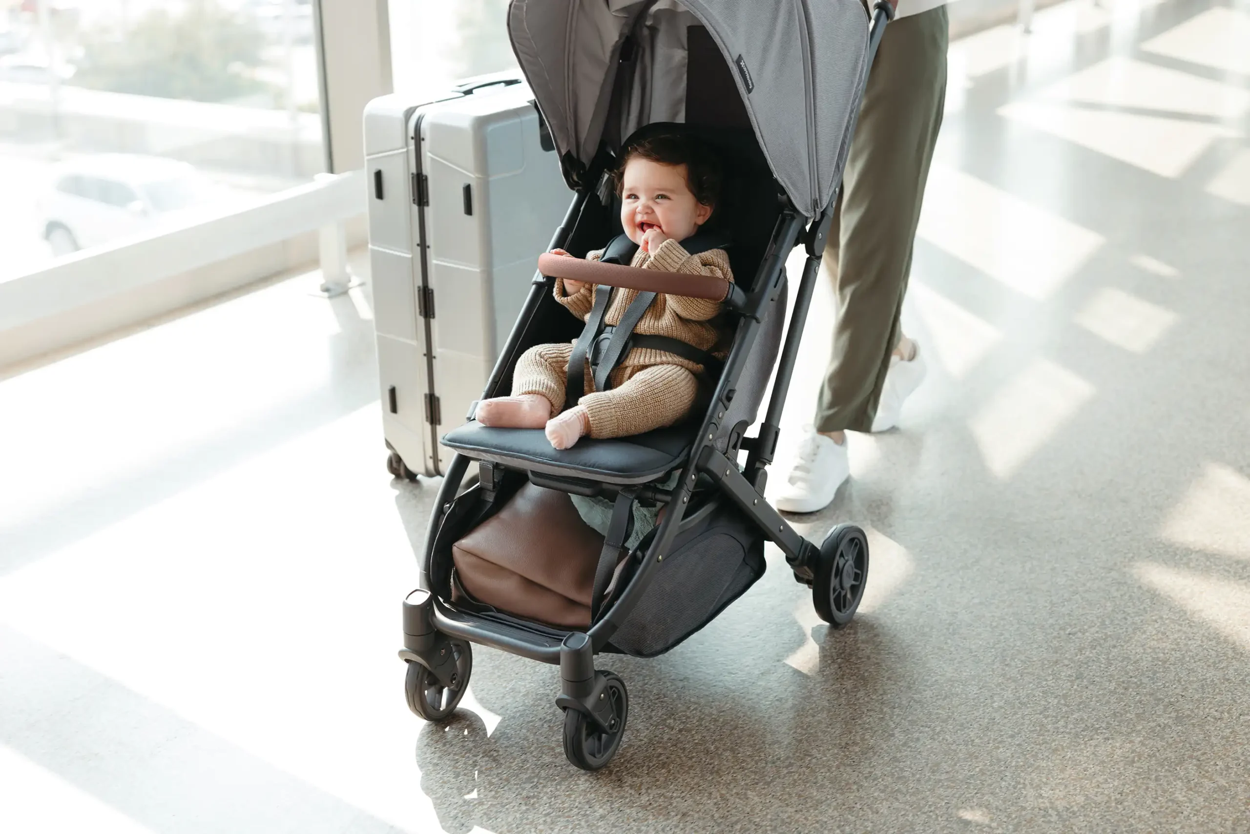A smiling toddler strolls comfortably in the Minu V2 while her mother's hands to wheel her luggage thanks to the stroller's large, easy access storage basket