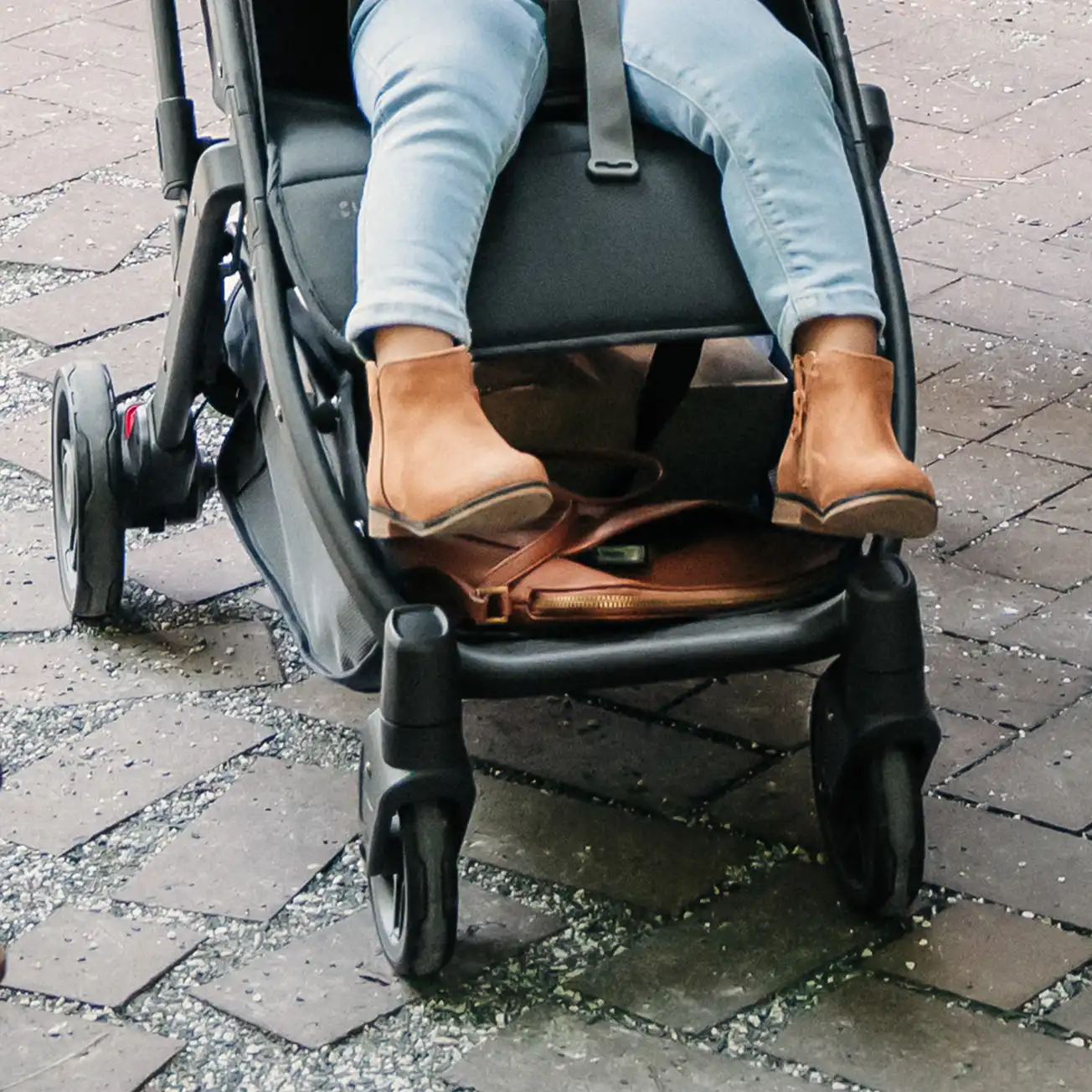 A toddler in a Minu V2 strolls through a brick city sidewalk with ease due to the stroller's front and rear spring-action suspension