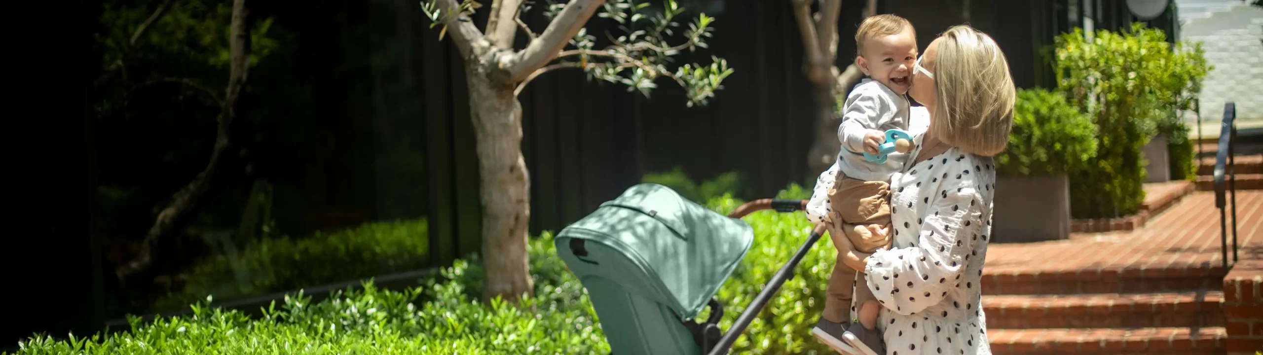 Mom carrying smiling infant and lovingly kisses baby's cheek while standing beside stroller