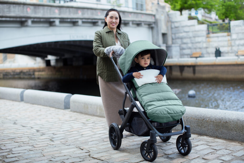 A woman pushing her child in an UPPAbaby V2, equipped with her UPPAbaby CozyHandmuffs and her UPPAbaby CozyGanoosh