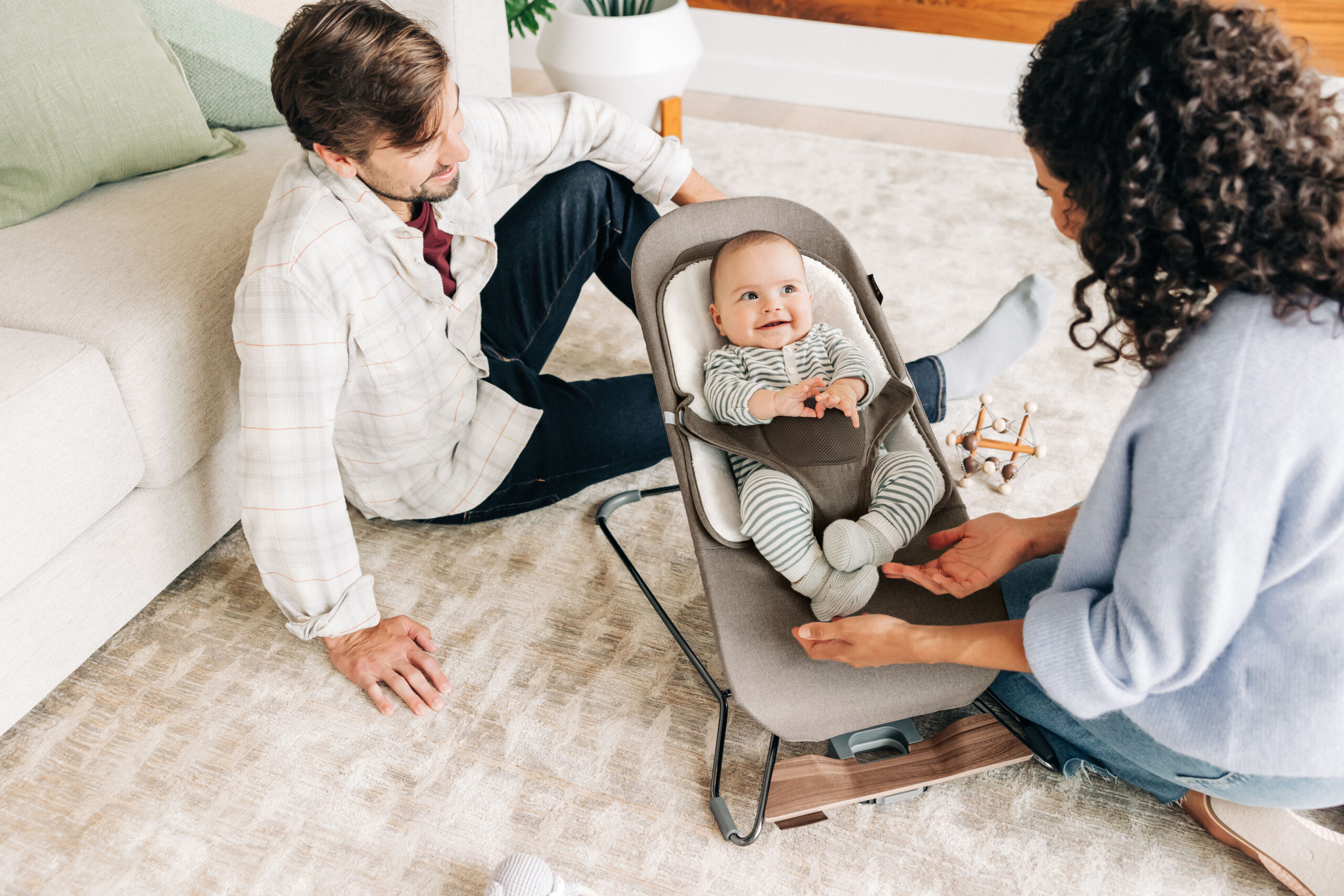 A candid moment with a baby smiling joyfully in the UPPAbaby Mira Bouncer placed on a living room rug, surrounded by relaxed parents engaging with the child, highlighting the bouncer's use in a family setting.