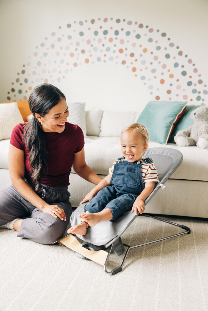 A baby lies comfortably in the UPPAbaby Mira Bouncer on a home rug, with a parent gently adjusting the seat angle, illustrating the bouncer’s ease of use and adjustable functionality.