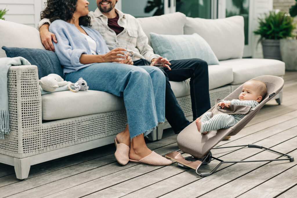 An UPPAbaby Mira Bouncer positioned on an outdoor wooden deck next to a cozy sofa where two parents are sitting and conversing, with their baby nearby in the bouncer, enjoying the outdoors.