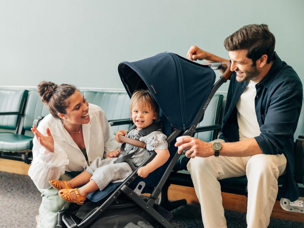 A mother and father sit with their child in a Noa Minu V2 travel stroller in an airport as they get ready to embark on an adventure