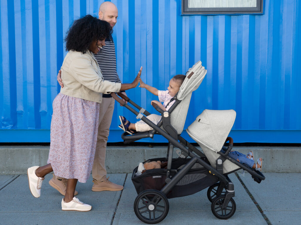 A mother and father happily stroll with a double Vista V2 stroller in Anthony fashion equipped with a RumbleSeat and both upper and lower adapters