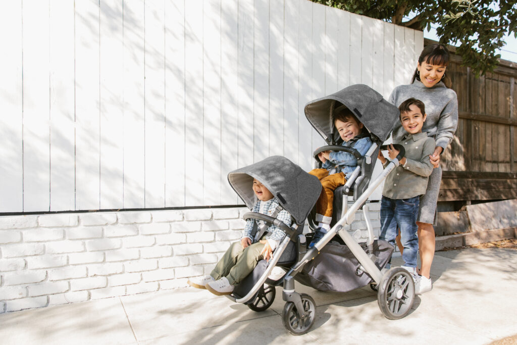 A smiling family using their Vista V2 stroller in a triple configuration, with the help of a RumbleSeat, upper and lower adapters, and a Piggyback ride along board