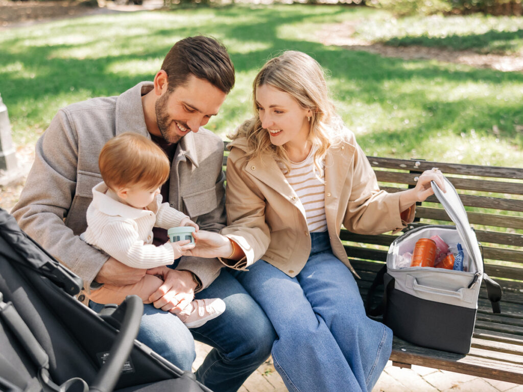 A family sits on a park bench enjoying some snacks and drinks kept cool in the Bevvy cooler after a stroll