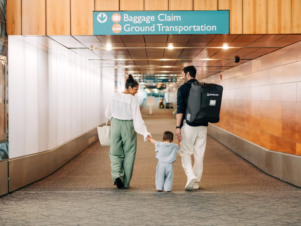 A man, woman, and child walk down a airport corridor after picking up their Minu V2 stroller (in a travel bag) from the claim