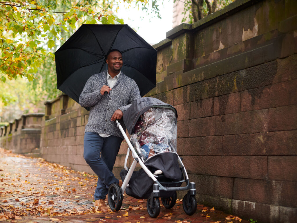 A man strolls with his child on a Fall day, who is staying dry in the Vista V2 stroller with the help of the protective Performance Rain Shield