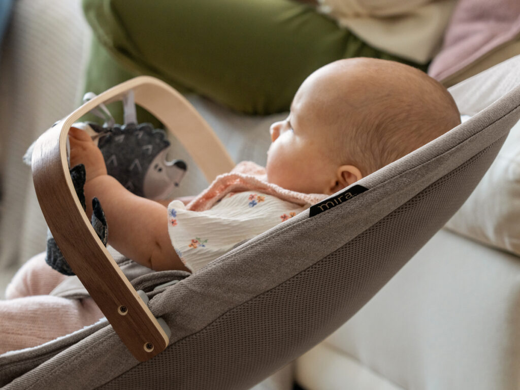 An infant comfortably bounces in the Mira bouncer, engaged with the Forest Fun toy bar. 