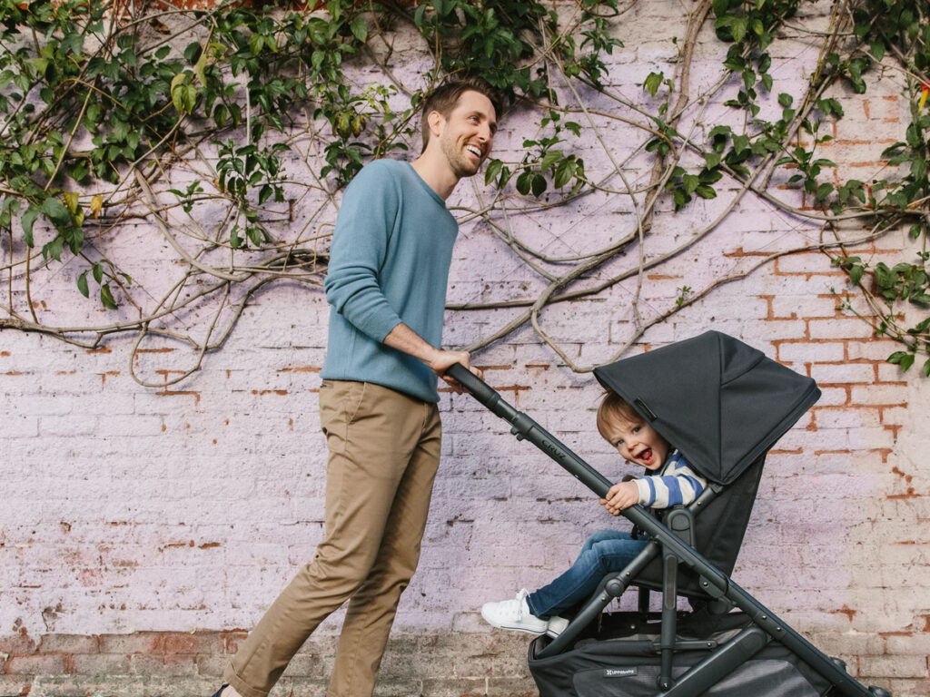 A smiling man walks with his Cruz stroller as his happy child peeks out at the world.