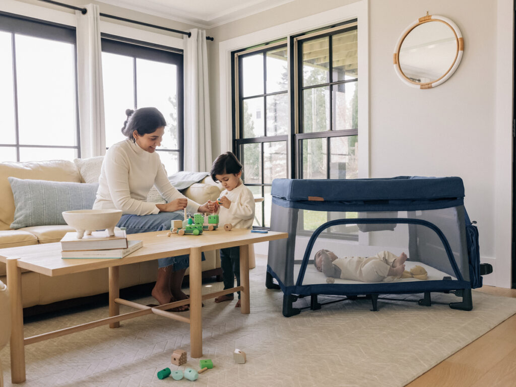 A baby lies comfortably in the Remi Playard, allowing a mother and toddler to play off to the side without worry