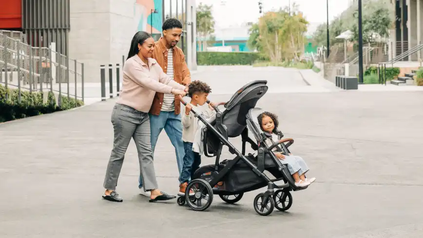 Mom and dad pushing their three children in Vista V3 with parent facing toddler seat, lower RumbleSeat and PiggyBack attachment